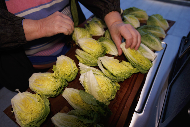 Zwei Hände ordnen mehrere Römersalat-Herzen auf einem dunklen Holzbrett an. Der Salat ist hellgrün und gelb. Im Hintergrund ist ein blass gestreiftes Oberteil sichtbar und links ein dunkler Ärmel. Teile einer Edelstahl-Oberfläche sind rechts im Bild.